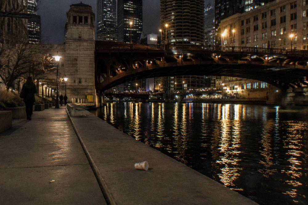 A dark Chicago riverfront walkway with the lights of downtown bridges reflected in the water