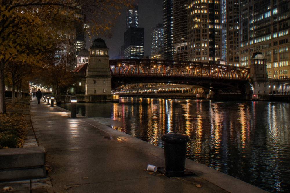 A wide sidewalk beside the Chicago River at night with bridge towers lit by street lamps