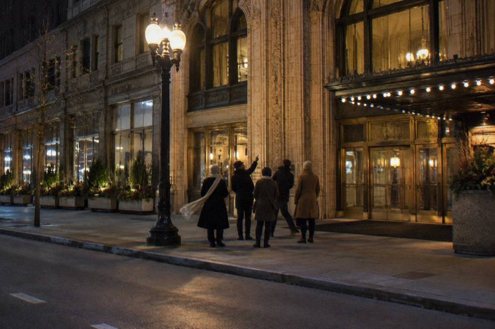A guided walking group paused on a Loop sidewalk at night under streetlights