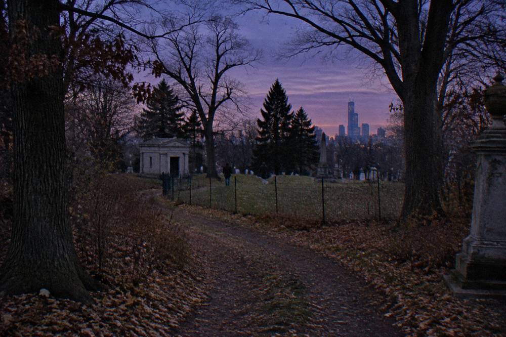 A narrow dirt path through dense woods leading toward a small fenced clearing in daylight