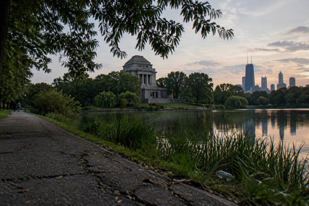 A tree-lined park path beside a lagoon at dusk with a stone building beyond the trees