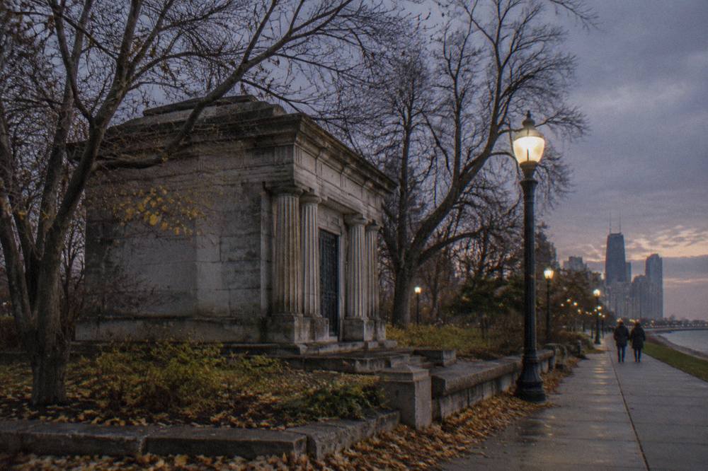 A Chicago street at dusk with streetlamps, bare trees and a darkening sky over the sidewalk