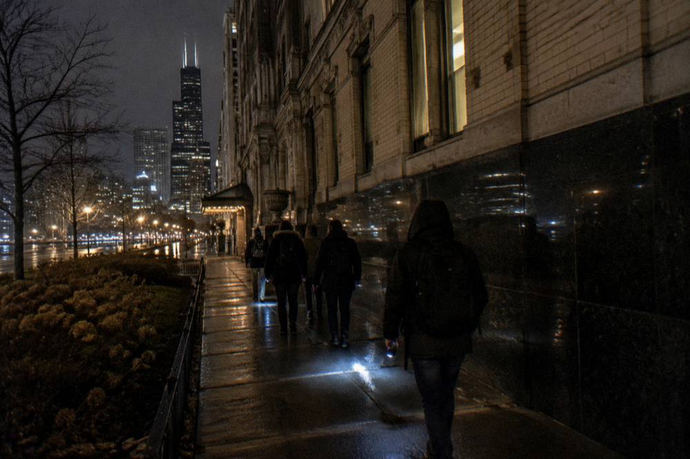A small group with flashlights walking a dark city sidewalk past old brick buildings