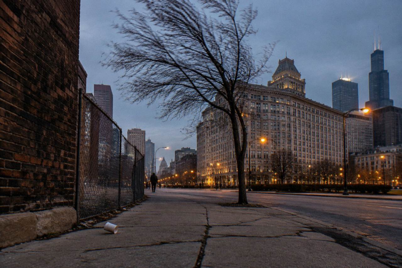An empty street corner in a South Side Chicago neighborhood at dusk with a low brick building behind a wire fence
