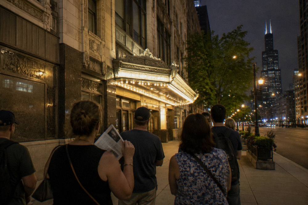 Evening sidewalk outside a lit Chicago Loop theater with a small tour group gathered