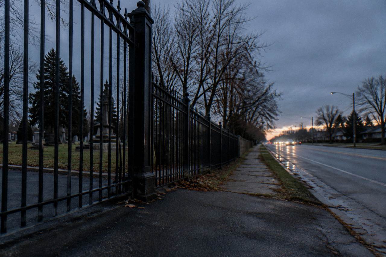 A tree-lined road curving past an old iron cemetery gate at dusk