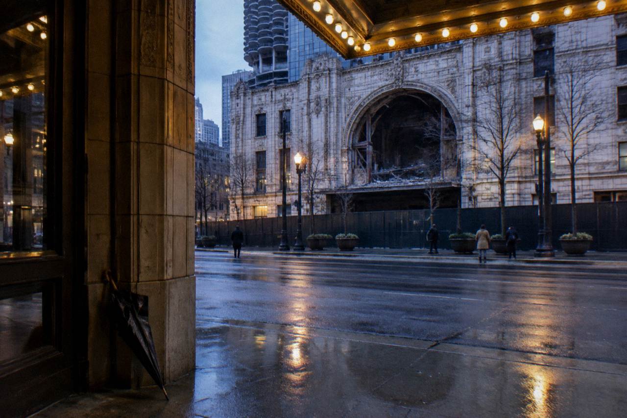 A lamplit Chicago sidewalk at dusk with old brick buildings and a lit theatre sign in the background